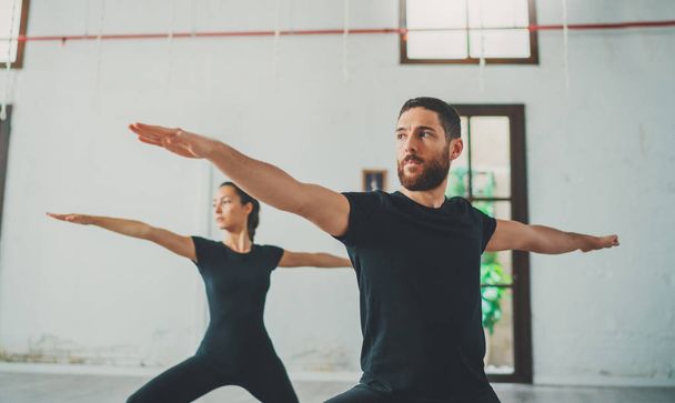 Young sportive man and woman are practicing yoga exercises in the studio. Couple of young sporty people practicing yoga lesson with partner - Фото, изображение