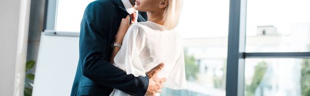 panoramic shot of businessman hugging blonde coworker while flirting in office  - Фото, изображение