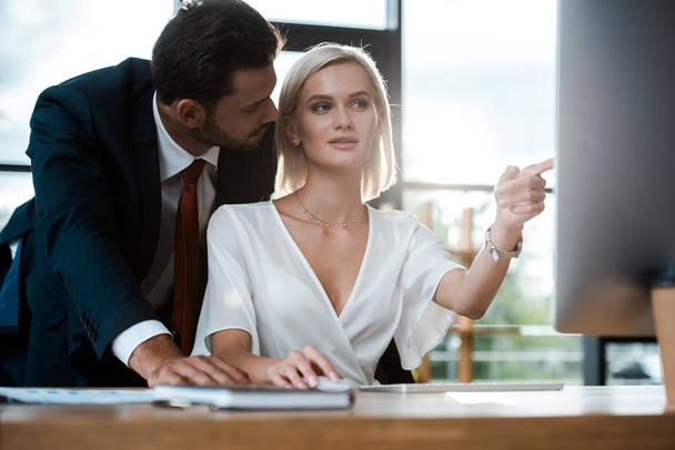 selective focus of bearded businessman looking at attractive girl pointing with finger at computer monitor  - Photo, Image