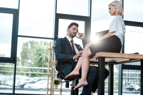 handsome man touching leg of attractive woman sitting on table in office  - Valokuva, kuva