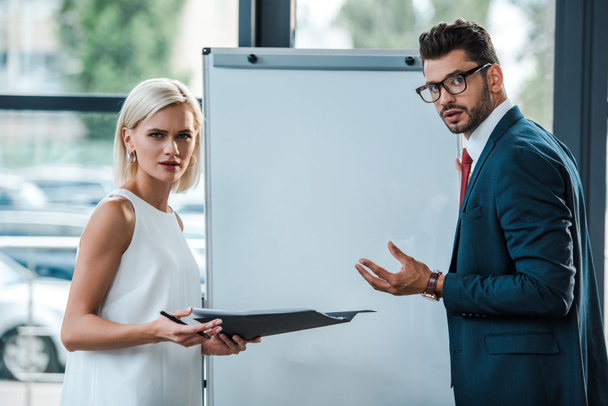 attractive blonde woman holding folder near businessman in glasses gesturing in office  - Photo, Image