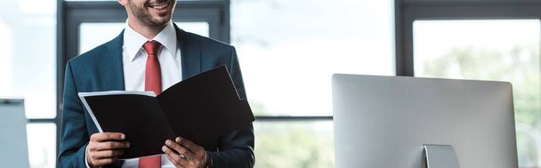 panoramic shot of cheerful man holding folder near computer monitor in office  - Photo, Image