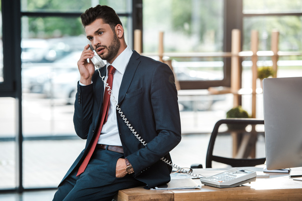 handsome bearded man talking on vintage phone and sitting on table with hand in pocket  - Photo, image