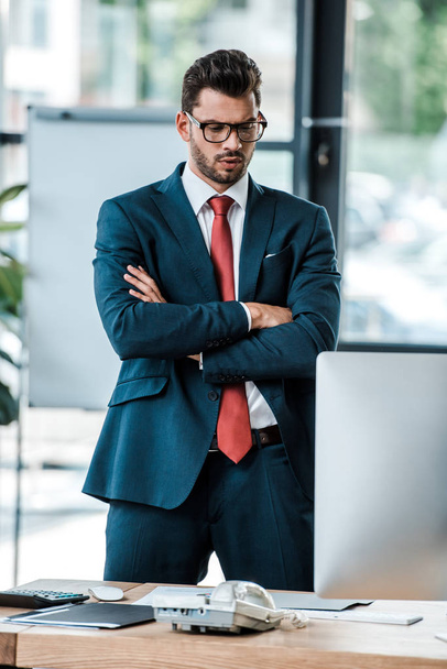 serious businessman in glasses standing with crossed arms and looking at computer monitor  - Foto, imagen