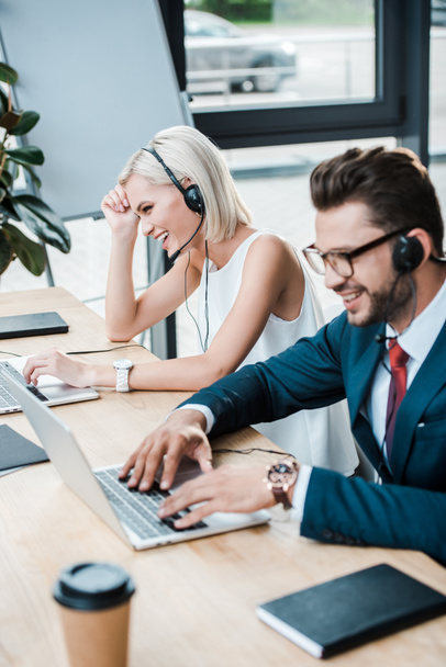 selectieve focus van gelukkige vrouw in headset in de buurt van vrolijke collega die laptop gebruikt tijdens het werken in Office  - Foto, afbeelding