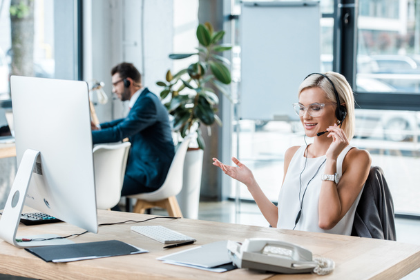 foco seletivo de mulher loira alegre em gesto headset enquanto fala perto colega de trabalho
  - Foto, Imagem
