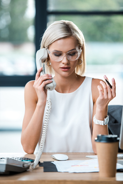 selective focus of businesswoman in glasses looking at documents and talking on retro phone near paper cup - 写真・画像
