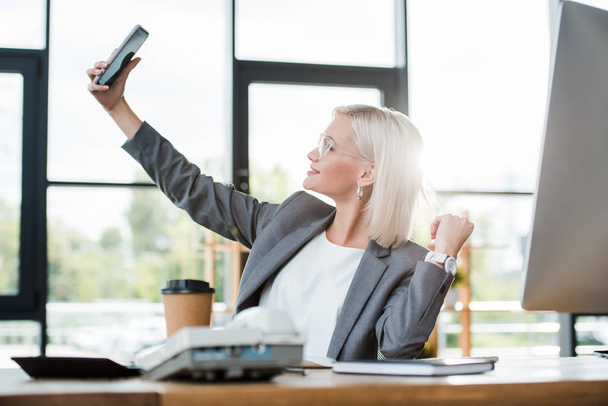 selective focus of cheerful businesswoman in glasses taking selfie in modern office  - Fotó, kép