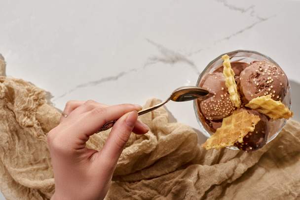 cropped view of woman holding soon near delicious chocolate ice cream in bowl with waffles and brown cloth on marble grey background - Fotoğraf, Görsel