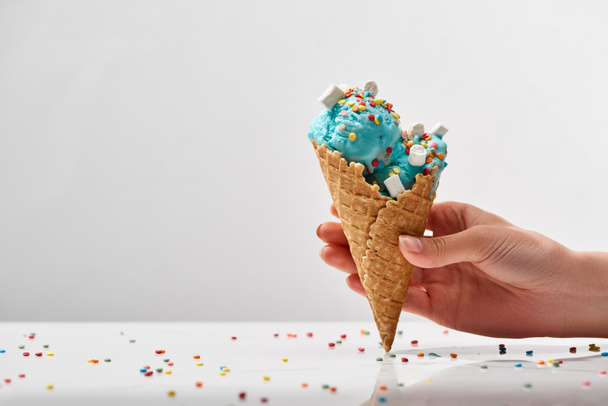 cropped view of woman holding delicious sweet blue ice cream with marshmallows and sprinkles in crispy waffle cone isolated on grey - Photo, Image