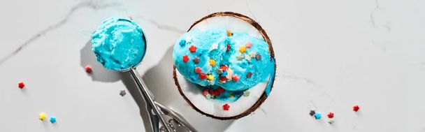 vista superior de delicioso helado azul con aspersiones sobre la mitad de coco y en cuchara de helado sobre fondo gris mármol, plano panorámico
 - Foto, Imagen