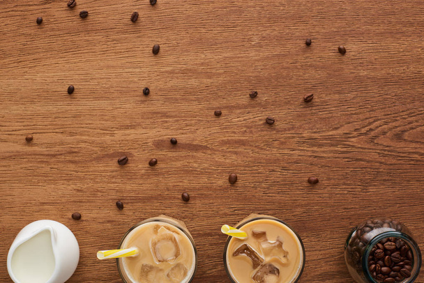top view of coffee in glasses with ice and straws, milk and coffee grains on wooden table with copy space - Photo, Image