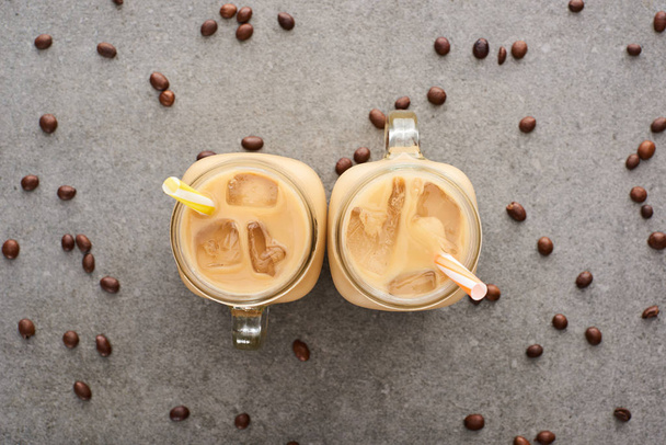 top view of ice coffee in glass jars with straws and coffee grains on grey background - Foto, afbeelding