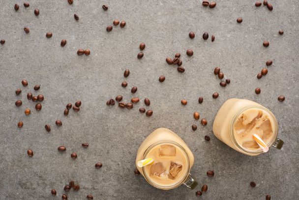 top view of ice coffee in glass jars with straws and scattered coffee grains on grey background - Valokuva, kuva