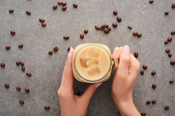 cropped view of woman holding glass jar with ice coffee and coffee grains on grey background - Foto, afbeelding