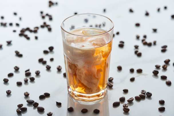 selectieve focus van ijskoffie mengen met melk in glas in de buurt van verspreide koffie korrels op grijze achtergrond - Foto, afbeelding