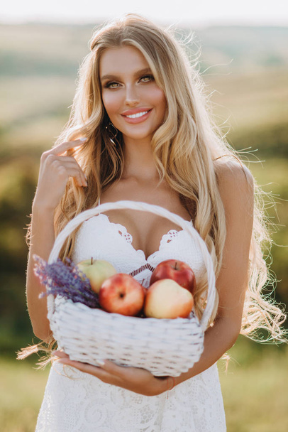 Beautiful sexy blonde girl in white dress posing in a field at sunset with a basket of fruit  - Zdjęcie, obraz