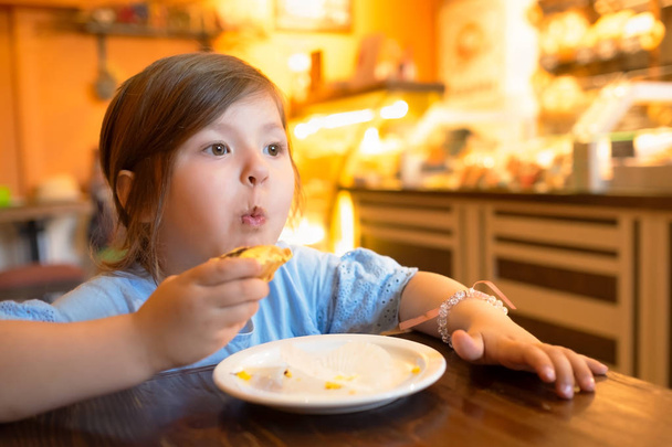 Χαρούμενο ωραίο μικρό κορίτσι κάθεται στο τραπέζι, ενώ τρώει γλυκό κέικ - Φωτογραφία, εικόνα