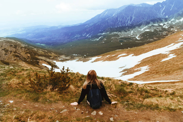 Νεαρή ξανθιά ταξιδεύει με ένα μπλε σακίδιο, κάθεται στην κορυφή ενός βουνού και απολαμβάνει καταπράσινο ορεινό τοπίο. Πίσω όψη - Φωτογραφία, εικόνα