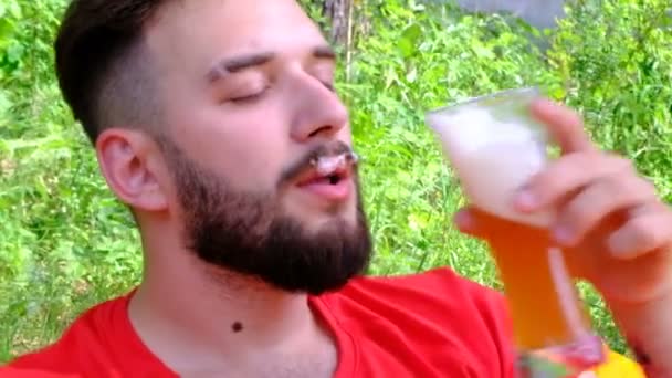De man in het rode t-shirt drinkt bier. Schuim op de snor. Een jonge man baard ligt op een oranje lucht Bank in een clearing in het bos. Close-up. Concept vakantie, picknick, reizen en lifestyle. - Video