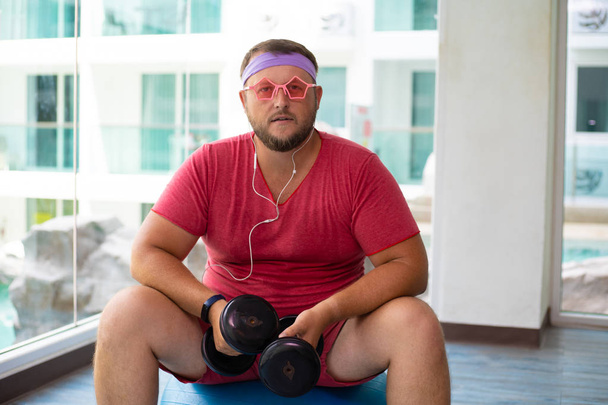 Смішний товстий чоловік в рожевих окулярах і в рожевій футболці займається гантелі на підходящому м'ячі в спортзалі, зображуючи дівчину
 - Фото, зображення