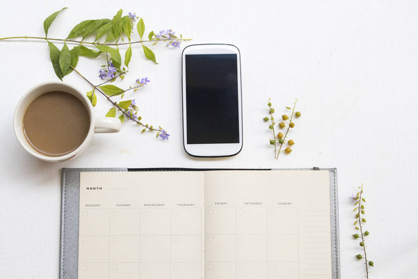 Notebook Planner z telefonem komórkowym do pracy i fioletowe kwiaty, gorący układ kawy płaski lay styl na białym tle na biurku biurowego  - Zdjęcie, obraz