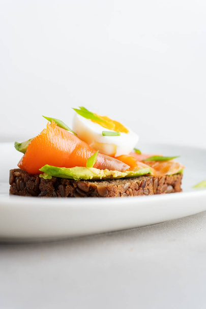 Sándwich saludable en tostadas integrales multigrano con aguacate, salmón, huevo y cebolla de primavera sobre fondo de plato blanco
 - Foto, imagen