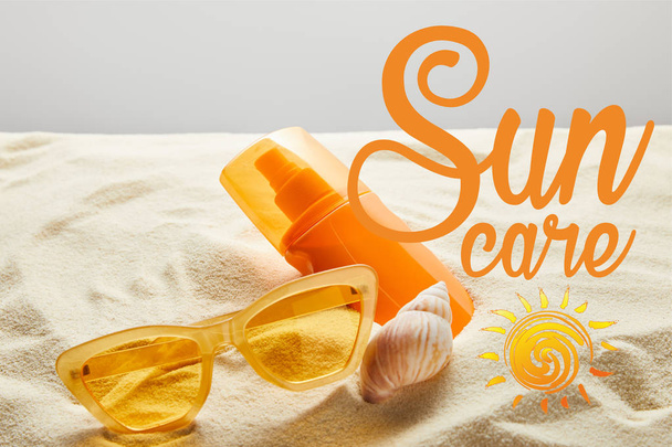 Gelbe stylische Sonnenbrille und Sonnencreme in orangefarbener Flasche auf Sand mit Muschel auf grauem Hintergrund mit Sonnenpflege-Schriftzug - Foto, Bild