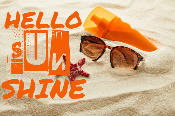 brown stylish sunglasses and sunscreen in orange bottle on sand with hello sunshine lettering - Фото, изображение