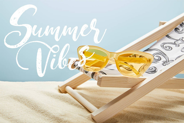 amarelo óculos de sol elegantes na cadeira deck na areia e fundo azul com vibrações de verão lettering
 - Foto, Imagem