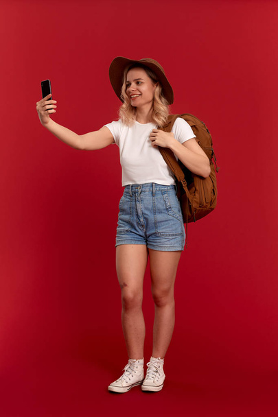 Portrait of a girl with curly blond hair dressed in a sundwon hat and white t-shirt standing on a red background making selfie with mobile phone. Model travels with orange backpack. - Fotó, kép
