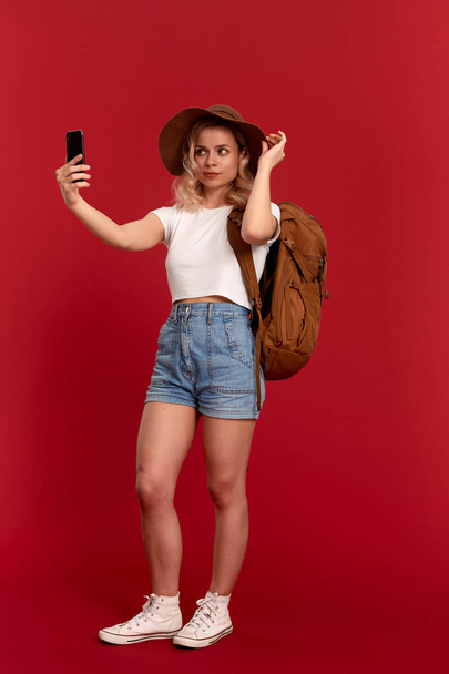 Porträt eines Mädchens mit blonden Locken, Sonnenhut und weißem T-Shirt, das vor rotem Hintergrund steht und ein Selfie mit dem Handy macht. Model reist mit orangefarbenem Rucksack. - Foto, Bild