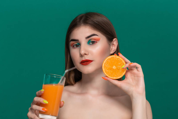 Closeup portrait of beautiful young woman with bright color make-up with orange juice holding half of an orange near the face. - Photo, Image