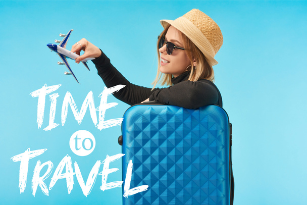 blonde happy girl in sunglasses and straw hat plating with toy plane near blue travel bag isolated on blue with time to travel illustration  - Fotoğraf, Görsel