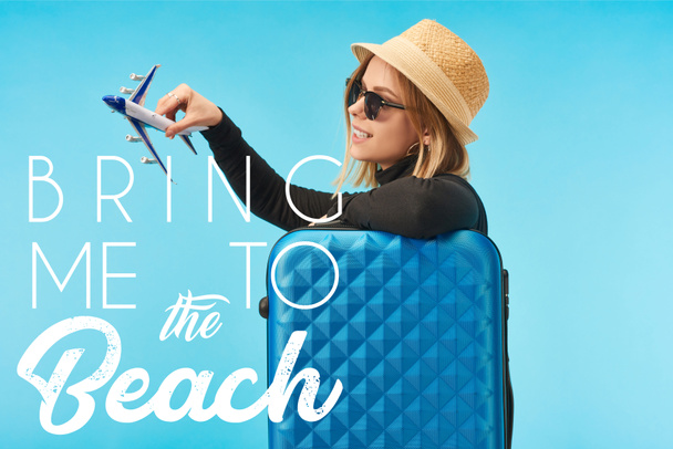 szőke boldog lány napszemüvegben és szalmakalapos galvanizálás játékgéppel közel kék utazási táska elszigetelt kék, hogy engem a strandon illusztráció - Fotó, kép