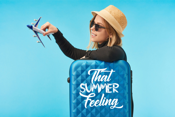 szőke boldog lány napszemüvegben és szalmakalapban plating játékgéppel közel kék utazási táska azzal a nyári érzés felirat elszigetelt kék - Fotó, kép