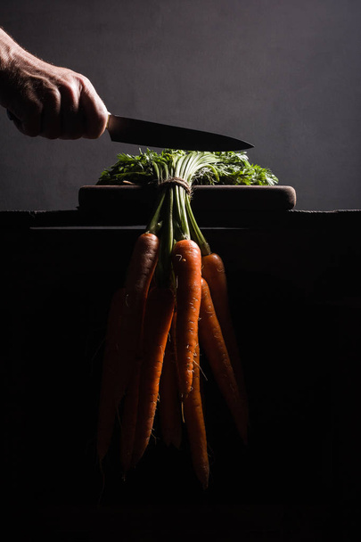 A hand holds a knife just before cutting the stems of the carrots hanging from the table - Fotoğraf, Görsel