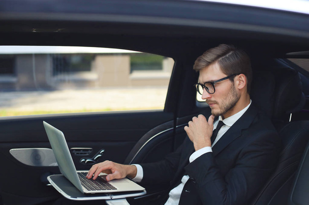 Thoughtful young businessman keeping hand on chin while sitting in the lux car and using his laptop. - Φωτογραφία, εικόνα