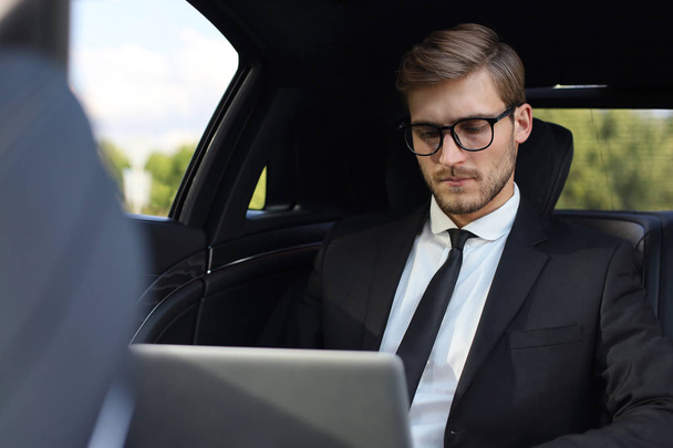Thoughtful confident businessman sitting in the luxe car and using his laptop. - Фото, изображение