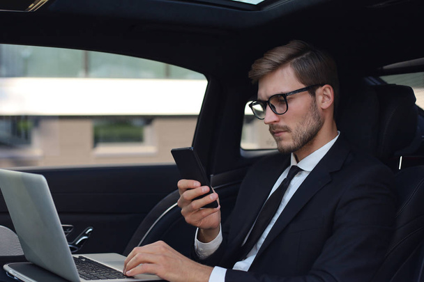 Handsome confident man in full suit looking at his smart phone while sitting in the car and using laptop. - Foto, afbeelding