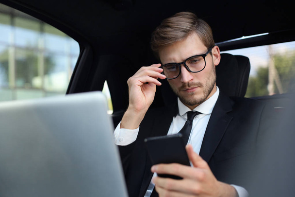 Handsome confident man in full suit looking at his smart phone while sitting in the car and using laptop. - Fotoğraf, Görsel