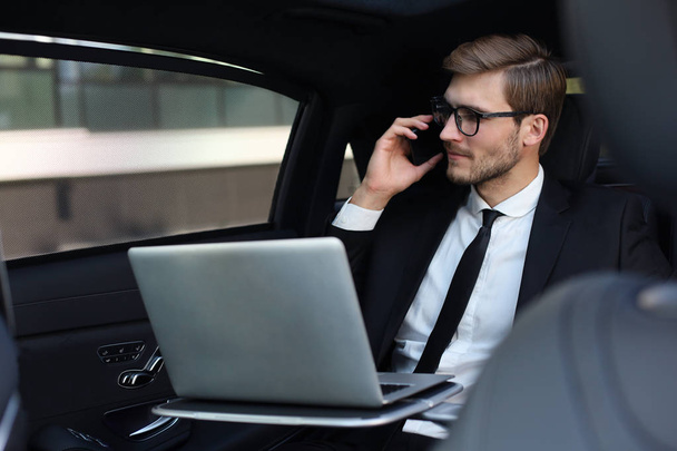 Takım elbiseli yakışıklı, akıllı telefonla konuşan ve arabada otururken dizüstü bilgisayar kullanarak çalışan yakışıklı kendine güvenen bir işadamı. - Fotoğraf, Görsel