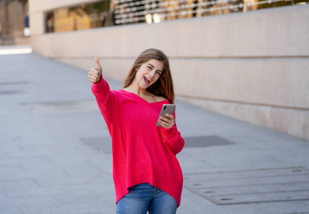Young attractive happy woman using mobile smart phone making thumb up sign outside in city street. Beautiful teenager wearing stylish clothes feeling carefree. Lifestyle and technology use concept. - Fotografie, Obrázek