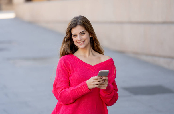 Pretty happy student woman chatting online on smart phone using social media apps outside city street. Beautiful teenager wearing stylish clothes feeling free. People, lifestyle and mobile use. - Photo, image