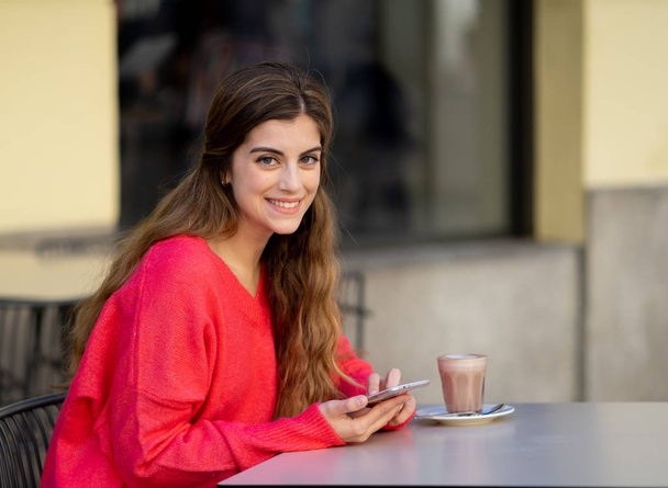 Mujer bastante joven buscando alegre bebiendo un café, enviando mensajes y charlando en su teléfono móvil inteligente fuera de una cafetería de la ciudad. Sentirse feliz y alegre. En estilo de vida y tecnología
. - Foto, Imagen