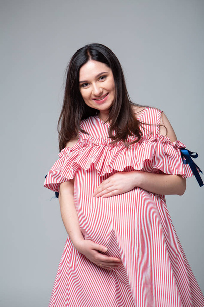 Pregnant brunette woman in a bright fashionable striped dress. Emotional studio photo on a white background. Professional light. Red and white stripe. Big belly. Last month of pregnancy. - Photo, Image
