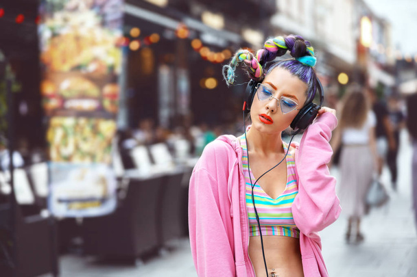Δροσερό φάνκι νεαρή γυναίκα με μοντέρνα γυαλιά ακούγοντας μουσική στα ακουστικά εξωτερική-χίππη κορίτσι με γυαλιά ηλίου και σκουλαρίκια απολαμβάνουν μουσική κύματα, εμφάνιση του δρόμου μόδας με κορίτσι εφήβων με τρελά μαλλιά - Φωτογραφία, εικόνα