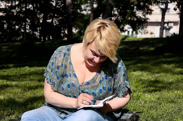 Woman and student sitting on grass, takes notes in notebook, learning and writes thoughts, writes book, she is preparing his final exams doing homework and thinking, writing something in his notebook. - Valokuva, kuva