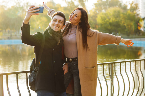 Happy young beautiful loving couple posing walking outdoors in park nature take selfie by mobile phone. - Foto, afbeelding