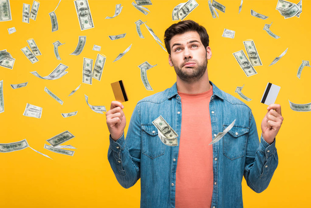 confused handsome man holding credit cards isolated on yellow with money rain illustration - Foto, Imagem